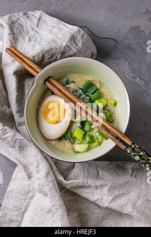 Weiße Schale mit asiatischen Stil Suppe mit Rührei, die Hälfte der marinierte Ei, Frühlingszwiebeln, Spinat serviert mit hölzernen Stäbchen Textil über graue textur Stockfoto