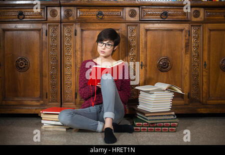 Junge Studentin mit Blick in die Kamera sitzen auf dem Boden mit vielen Büchern rund um müden Gesichtsausdruck Stockfoto