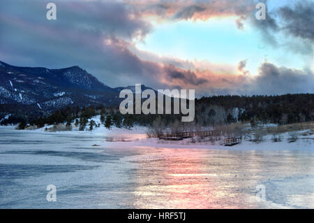 Majestic parting of the clouds during a stormy sunset filters pink and blue into a reflection on this frozen lake.  Wooden pier is backlighted. Stockfoto
