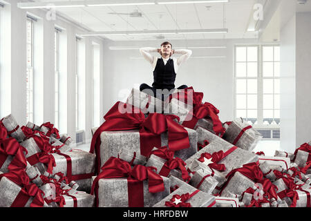 Junger Geschäftsmann auf Stapel von Geschenk-Boxen sitzen. Mixed-media Stockfoto