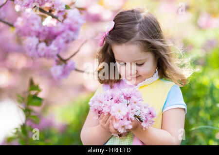 Glückliche Mädchen spielen unter blühenden Kirschbaum mit rosa Blüten. Kind hält Sakura Blüte. Sommer Spaß für Familien mit Kindern im Freien in einem sein Stockfoto