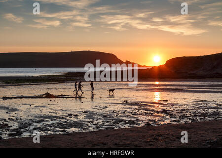 Eine Familie mit Hund in der Silhouette bei Sonnenuntergang an einem Strand in der Daymer Bay in Cornwall wandern.  Die untergehende Sonne spiegelt sich auf dem nassen Sand und das Meer Stockfoto