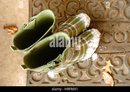 Grüne Damen Gummistiefel oder Gummistiefel, mit Schnee auf den Zehen gemustert. Auf eine willkommene Matte außerhalb eines Hauses. Stockfoto