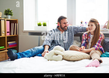 Teenager-Tochter in kariertes Hemd sitzt auf dem Bett neben ihrem Vater und erzählt ihm Probleme, er ihr aufmerksam zuhören und beraten Stockfoto