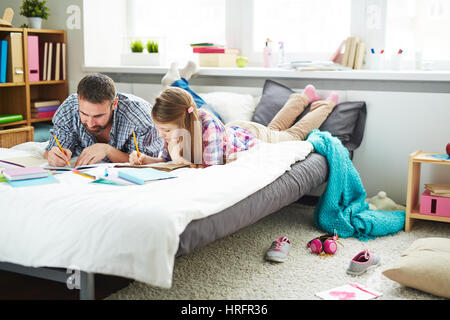 Konzentrierte Mann und seiner kleinen Tochter am Bett liegen und zeichnen von Bildern mit Interesse Stockfoto