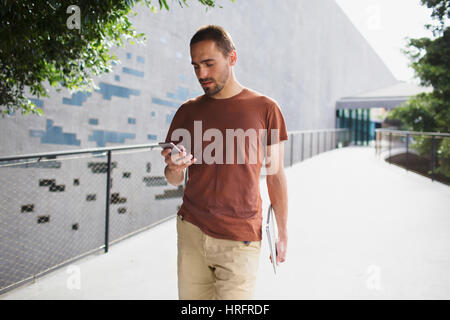 Young bärtiger Mann in Freizeitkleidung geht entlang dem Pfad und SMS Nachricht auf seinem smartphone Stockfoto