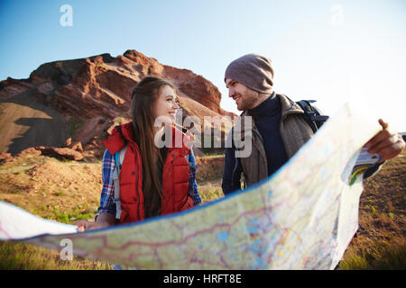 Junge fröhliche paar von Abenteurern, die riesige Karte halten und und sahen einander lächelnd auf Wanderweg in Bergen von hellem Sonnenlicht beleuchtet Stockfoto
