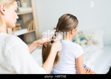 Junge Frau sorgfältig Flechten ihre Töchter schöne lange Haare sitzen im sonnendurchfluteten Kinderschlafzimmer Stockfoto