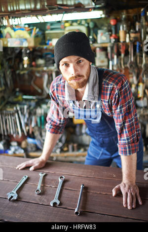 Porträt der schönen bärtigen Mechaniker tragen einheitliche Overall stützte sich auf Tisch und Blick selbstbewusst in die Kamera vor Hintergrund der Regale füllen Stockfoto