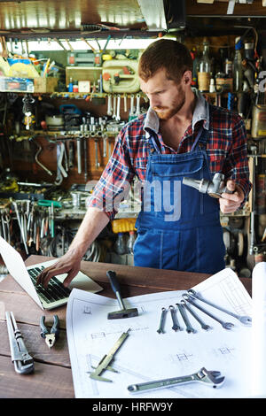 Porträt der schönen bärtigen Mechaniker tragen einheitliche Overall beschäftigt in Garage arbeiten, Montage von Teilen, die mit Werkzeugen und Baupläne beim Blick nach unten Stockfoto