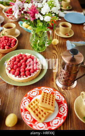 High-Angle Ansicht des frisch gebackenen Himbeer-Tarte, hausgemachte Waffeln, Sommerblumen, französische Presse und Tee Tassen platziert auf Holztisch Stockfoto