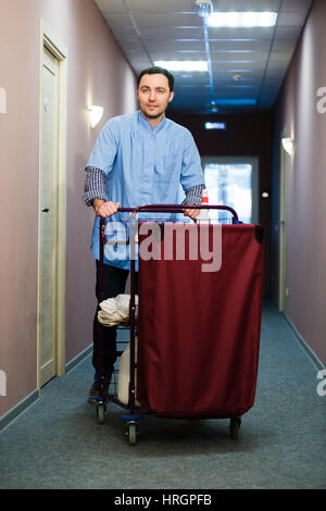 Junger Mann schob einen Haushalt Wagen beladen mit saubere Handtücher, Wäsche und Reinigungsgeräte in einem Hotel, als er das Zimmer services Stockfoto