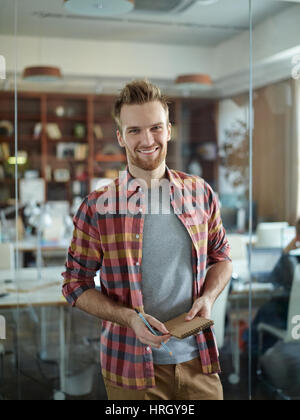Porträt von fröhlich gut aussehender Mann tragen bunt Kariert Hemd lächelnd zu Kamera stehend durch Glaswand im modernen Büro Stockfoto