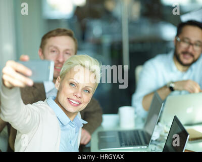 Porträt von mehreren Geschäftsleuten sitzt am Besprechungstisch in hellen modernen Konferenz-Zimmer und unter Selfie, Fokus auf blonde junge Frau trägt cre Stockfoto
