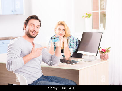 Getönten fröhliche junge bärtigen Lächeln auf den Lippen und zeigt zur Kamera Kreditkarte beim Sitzen vor dem Computer. Glücklich blonde Dame auf Hintergrund. Stockfoto
