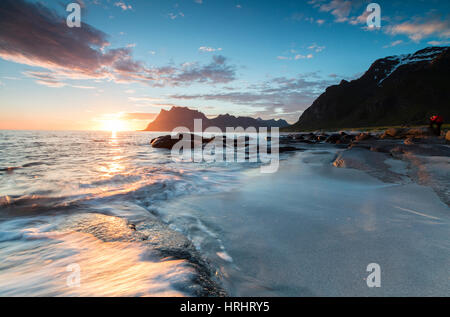 Rosa Wolken und Mitternachtssonne spiegelt sich auf den Wellen des blauen Meeres, umrahmt von felsigen Gipfeln, Uttakleiv, Lofoten Inseln, Norwegen Stockfoto