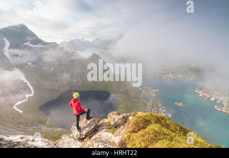 Wanderer am Gipfel bewundert, der blaue See und Meer umrahmen das Dorf, Reinebringen, Moskenesoya, Lofoten Inseln, Norwegen Stockfoto