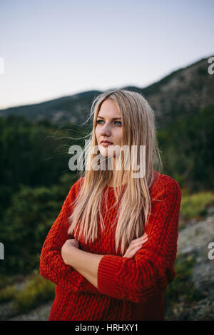 Porträt von eine attraktive junge blonde Frau im roten Pullover mit verschränkten Armen im Freien auf dem Hintergrund der Berge und Wald Stockfoto