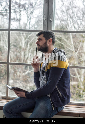 Porträt eines lässig bärtige Hipster denken, halten Sie Stift und Notizen im Heft oder Notepad zu Hause. Große Fenster Backdround. Handschrift Stockfoto