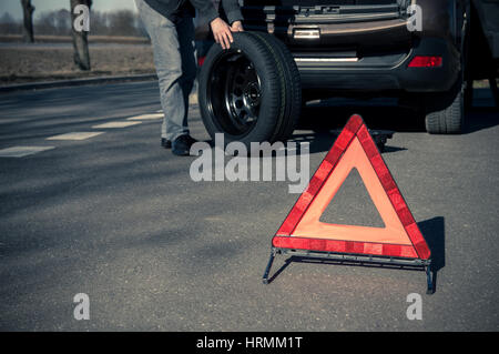 Warndreieck steht auf der asphaltierten Straße und Mann hält das Reserverad auf dem Hintergrund Stockfoto