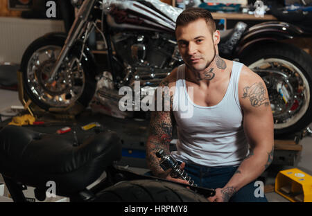 Richtige Detail. Brutale junge Biker mit Tattoo Jeans und weißes T-shirt auf dem Hintergrund der großen Motorrad sitzend und Blick in die Kamera Stockfoto