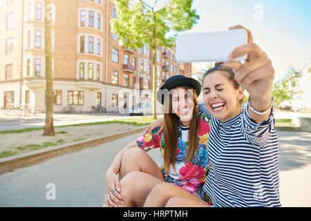 Ehrliche Bild einer lachenden jungen Frau, die unter ein Selbstporträt mit trendigen jungen Freundin, wie sie zusammen auf dem Bordstein urban ruhig sitzen Stockfoto