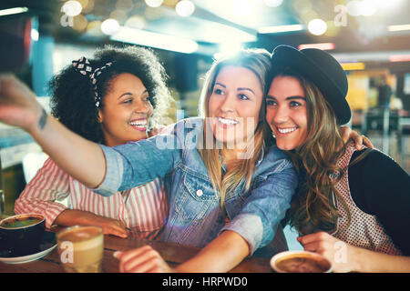 Drei lustige junge Frauen posieren für ein Selfie mit großen glücklichen Lächeln, während sie mit Kaffee in einem Bistro entspannen Stockfoto