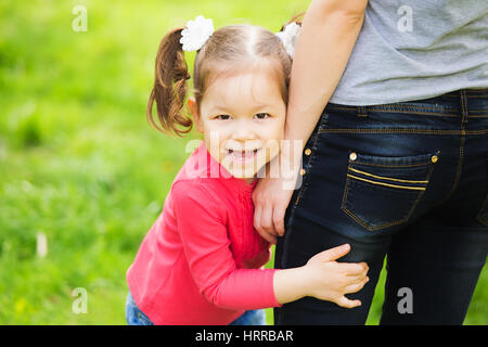 Closeup Portrait glückliche Familie. Kleine lustige Mädchen von 4 Jahren mit ihren Eltern im Stadtpark Frühling draußen spielen. Baby umarmt Hüften der Mutter und Stockfoto