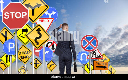Geschäftsmann ist aufgrund vieler Verkehrszeichen verwirrt, versucht, die richtige Richtung für das weitere Vorgehen entscheiden. Am Himmelshintergrund. Stockfoto