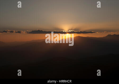 Untergehende Sonne hinter den Wolken und eine Silhouette von Bergen bei Einbruch der Dunkelheit in China. Stockfoto