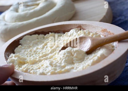 Geschmolzener Käse vorbereitet für den Käsekuchen in der Holzschale mit Löffel und einer weiblichen Hand gehalten. Auf dem Hintergrund sichtbar gerollte Käse Kuchen bereit f Stockfoto