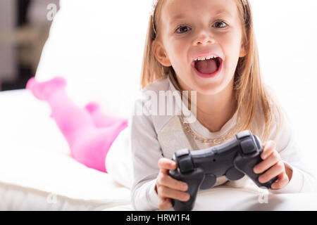 Aufgeregt, attraktive junge blondes Mädchen jubeln und Lachen mit Videospielen mit einem Controller in ihren Händen, wie sie auf ihrem Bett entspannt Stockfoto