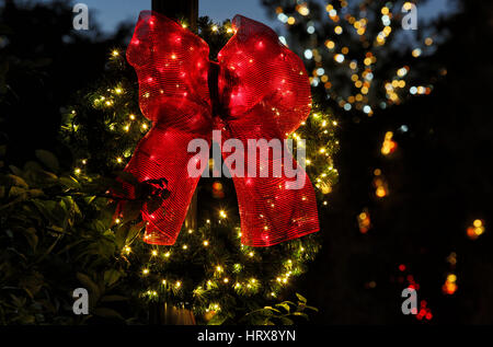 Ein beleuchteter immergrünen Adventkranz, mit einer großen roten Schleife, die auf einem dunklen Hintergrund mit Weihnachtsbeleuchtung im Hintergrund angezeigt. Stockfoto