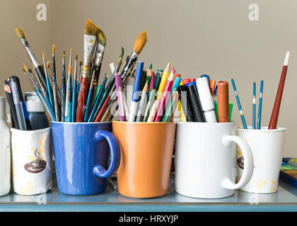 Nahaufnahme der verschiedenen verwendeten Pinsel, geschärften Buntstifte, Kugelschreiber und Marker auf farbige Tassen über blauer Tisch und Beige Hintergrund Stockfoto