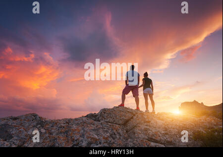 Silhouette von fröhlichen Menschen auf dem Berg gegen bunte Himmel bei Sonnenuntergang. Landschaft mit Silhouetten von stehender Mann und Frau mit Hand in Hand Stockfoto