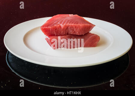 Frischer Thunfisch-Steak auf einem weißen Teller auf dem Tisch als schwarzer Hintergrund - frische rohe Scheiben. Korn ist die Textur der Glastisch. Stockfoto