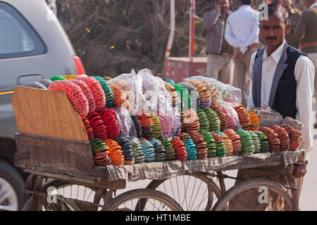 Eine nicht identifizierte Händler Räder seinen Karren der bunte Armbänder und Kopf Ringe für Verkauf zu einer nahe gelegenen Straße Tonhöhe in Varanasi, Indien Stockfoto