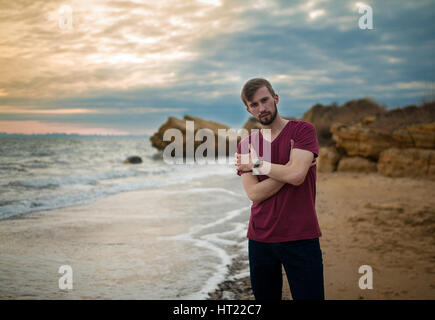 Mann im t-Shirt am Winter-Strand bei Sonnenuntergang. Wolken bei Sonnenuntergang Himmelshintergrund und Felsen sind von hinten sichtbar. Stockfoto