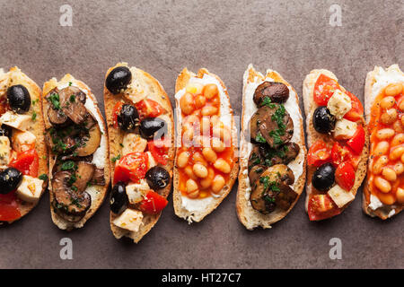 Große Auswahl an kleinen Bruschetta mit Caprese Salat, weißen Bohnen auf dem Grill mit Tomatensauce und gebratene Pilze mit Frischkäse, serviert auf graue Rückseite Stockfoto