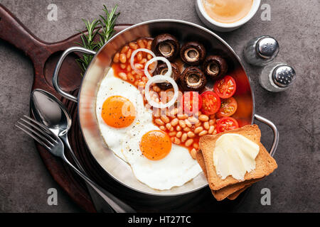 Frühstück mit Spiegeleiern in gusseisernen Pfanne, gebrochene Ei, Tomaten, auf Schneidebrett über graue Stein Hintergrund. Draufsicht mit Textfreiraum Stockfoto