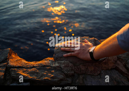 Schuss von Herren linke Hand auf einem Felsen, trägt eine einfache Ehering und eine Uhr hautnah. Meer und Sonnenuntergang Lichtspuren auf dem Hintergrund. Stockfoto