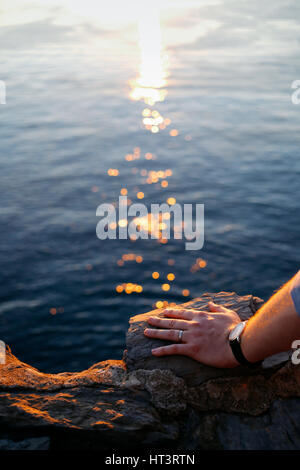 Schuss von Herren linke Hand auf einem Felsen, trägt eine einfache Ehering und eine Uhr hautnah. Meer, Sonnenuntergang und leichte Wanderwege auf dem Hintergrund. Stockfoto