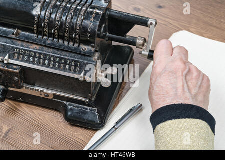 Alte faltige Hand dreht den Griff Retro-Rechenmaschine liegen auf dem Tisch neben einem Blatt Papier und einen Stift, Nahaufnahme Stockfoto