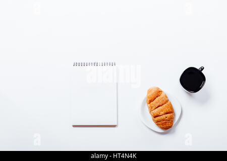 Croissant auf eine Untertasse, eine schwarze Tasse Kaffee und Notizbuch auf einem weißen Hintergrund, Ansicht von oben Stockfoto