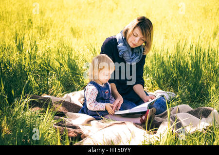 Porträt der glückliche Familie von zwei Personen im Urlaub. Junge Mutter und Töchterchen Buch sitzen auf der Picknickdecke in langen grünen Rasen. Alter von c Stockfoto