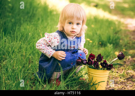 Porträt von glücklich kleine blonde Mädchen sitzen in der Nähe von gelben Eimer mit Tulpe Blumen. Süße lustige Kind spielt im sonnigen Frühlingsgarten in Landschaft. Alter o Stockfoto