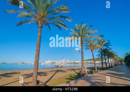 Vormittag sieht Sonne, Strand in der Nähe der Stadt, entlang einer verlassenen Segelboot.  Reihen von Palmen säumen den Strand, sonnigen Tag entlang der Wasserkante auf Ibiza. Stockfoto