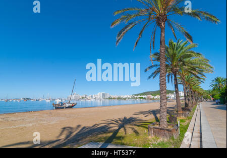 Vormittag sieht Sonne Strand entlang eines verlassenen Segelboot.  Reihen von Palmen säumen Wasserrand in Ibiza, Balearen St Antoni de Portmany, Spanien. Stockfoto
