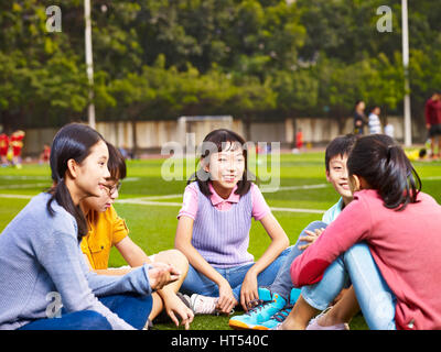 Gruppe von asiatischen Grundschule jungen und Mädchen sitzen und plaudern auf dem Spielplatz Rasen Stockfoto