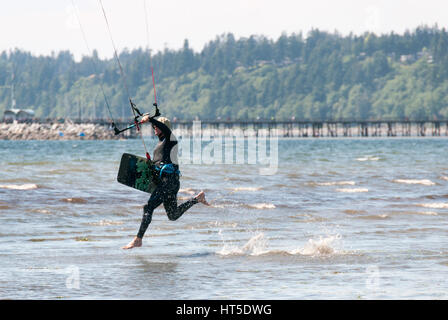 Mann läuft in der Brandung auf einem Strand Kitesurfen mit Brett in der hand einen Neoprenanzug tragen. Stockfoto